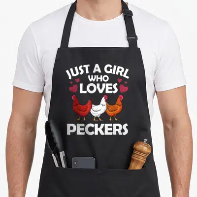 Flock chicken apron - Three cartoon chickens in white, red, and brown under Just A Girl Who Loves Peckers text — shown on a black cotton canvas apron against a white background