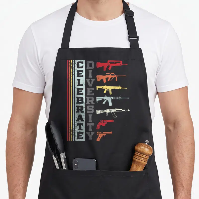 Different gun apron - Vertical CELEBRATE DIVERSITY text beside seven stacked colored gun silhouettes with distressed — shown on a black cotton canvas apron against a white background