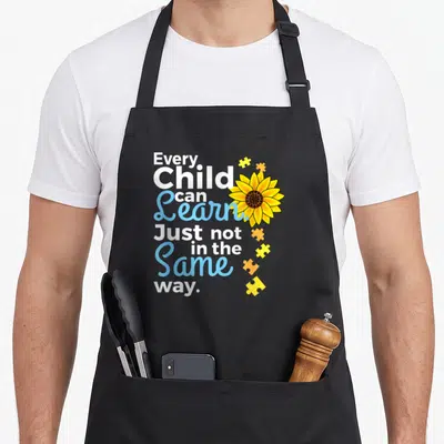 Special teacher art apron - Stacked text Every Child Can Learn with yellow sunflower and puzzle pieces — shown on a black cotton canvas apron against a white background