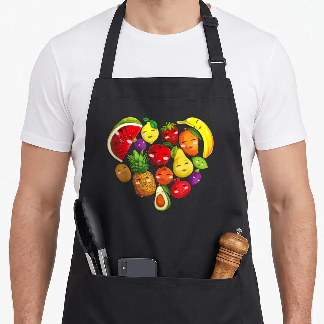 Collage vegetable apron - Heart shape formed by smiling cartoon fruits in red yellow green orange purple — shown on a black cotton canvas apron against a white background