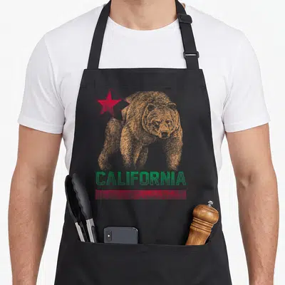 California republic bear apron - Orange bear illustration with red star and green CALIFORNIA text on distressed — shown on a black cotton canvas apron against a white background