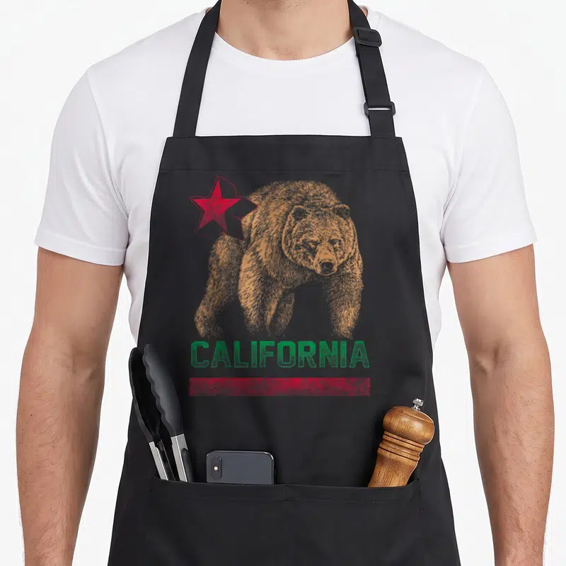California republic bear apron - Orange bear illustration with red star and green CALIFORNIA text on distressed — shown on a black cotton canvas apron against a white background