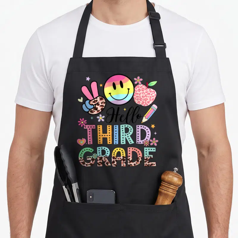 3rd grade teacher apron - Hello THIRD GRADE text with leopard apple, rainbow smiley, peace sign, pencil, flowers — shown on a black cotton canvas apron against a white background