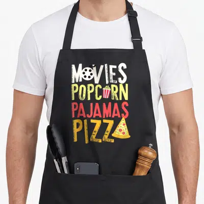 Movy pizza apron - Stacked text MOVIES POPCORN PAJAMAS PIZZA with film reel and pizza slice graphics — shown on a black cotton canvas apron against a white background