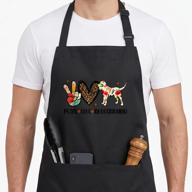 Peace bloodhound dog apron - Cartoon peace sign, leopard heart, and floral bloodhound with text PEACE LOVE BLOODHOUND — shown on a black cotton canvas apron against a white background