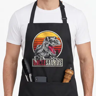 Uncle unclesaurus dinosaur apron - Grey T-Rex with sunglasses and striped sunset behind UNCLESADRUS text box — shown on a black cotton canvas apron against a white background