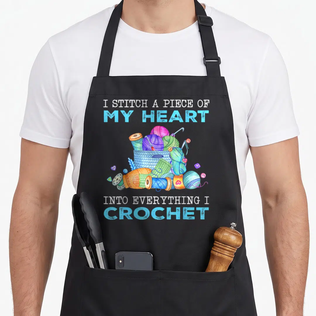 Funny stitch crochet apron - I STITCH A PIECE OF MY HEART INTO EVERYTHING I CROCHET text above and below a basket — shown on a black cotton canvas apron against a white background