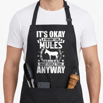 Okay mule apron - White mule silhouette inside horseshoe with stacked sarcastic text — shown on a black cotton canvas apron against a white background
