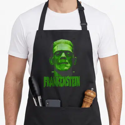 Universal frankenstein apron - Realistic green and grey Frankenstein monster head above dripping text — shown on a black cotton canvas apron against a white background