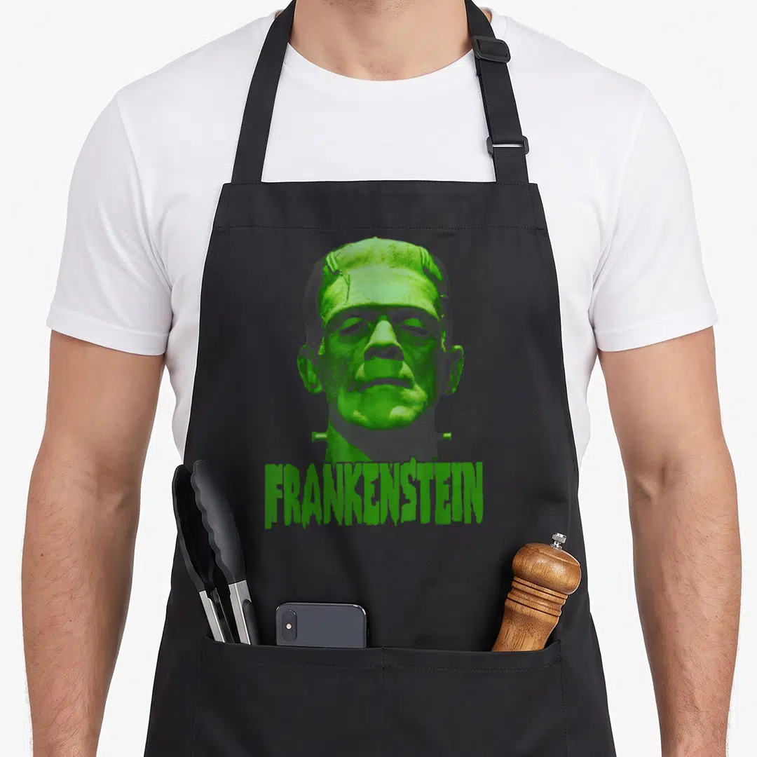 Universal frankenstein apron - Realistic green and grey Frankenstein monster head above dripping text — shown on a black cotton canvas apron against a white background
