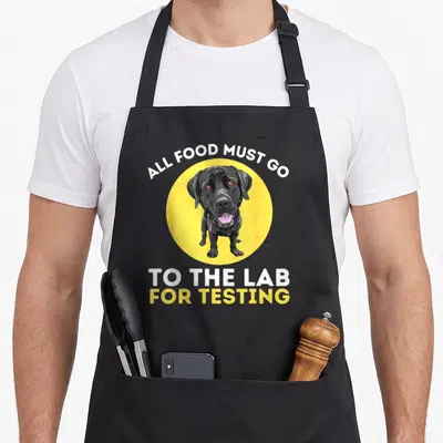 Testing labrador apron - Black cartoon dog in yellow circle with text All Food Must Go To The Lab For Testing — shown on a black cotton canvas apron against a white background
