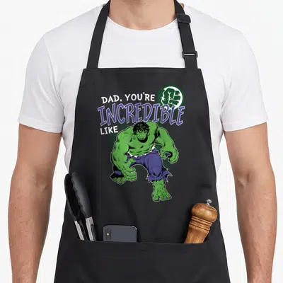 Retro incredible father day apron - Green cartoon character crouching below white and purple text reading DAD YOU'RE — shown on a black cotton canvas apron against a white background