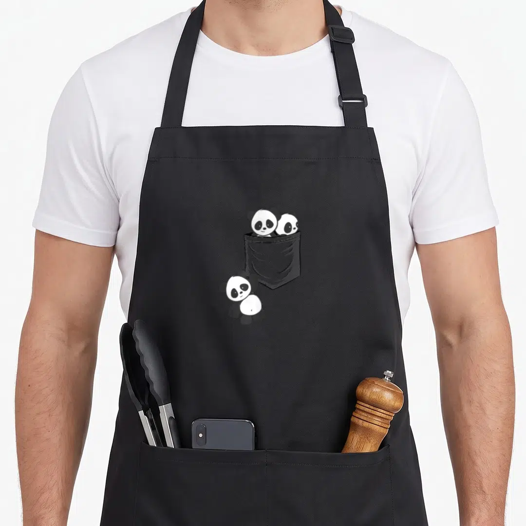 Baby kawaii apron - Three cartoon pandas in black and white, two in a grey pocket, one standing below — shown on a black cotton canvas apron against a white background