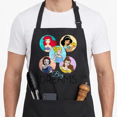 Sparkle princess apron - Five circular princess portraits above Big DREAMS text in blue and black — shown on a black cotton canvas apron against a white background