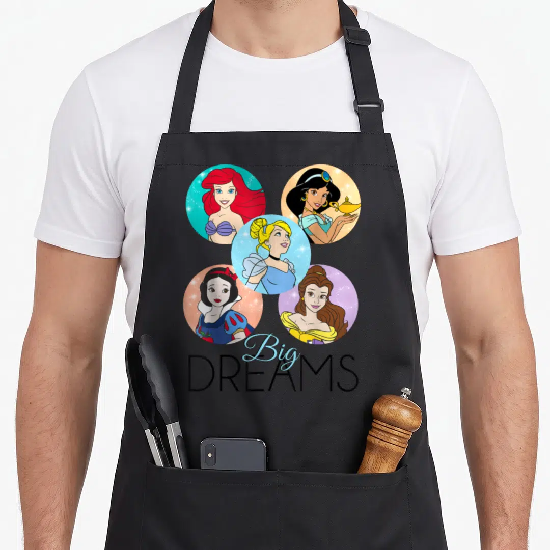 Sparkle princess apron - Five circular princess portraits above Big DREAMS text in blue and black — shown on a black cotton canvas apron against a white background