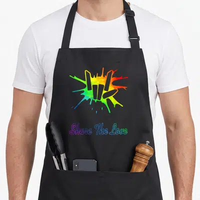 Share shared birthday apron - Rainbow watercolor splash above Share The Love text in gradient script — shown on a black cotton canvas apron against a white background