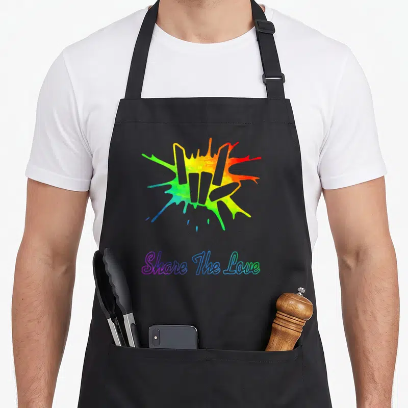 Share shared birthday apron - Rainbow watercolor splash above Share The Love text in gradient script — shown on a black cotton canvas apron against a white background
