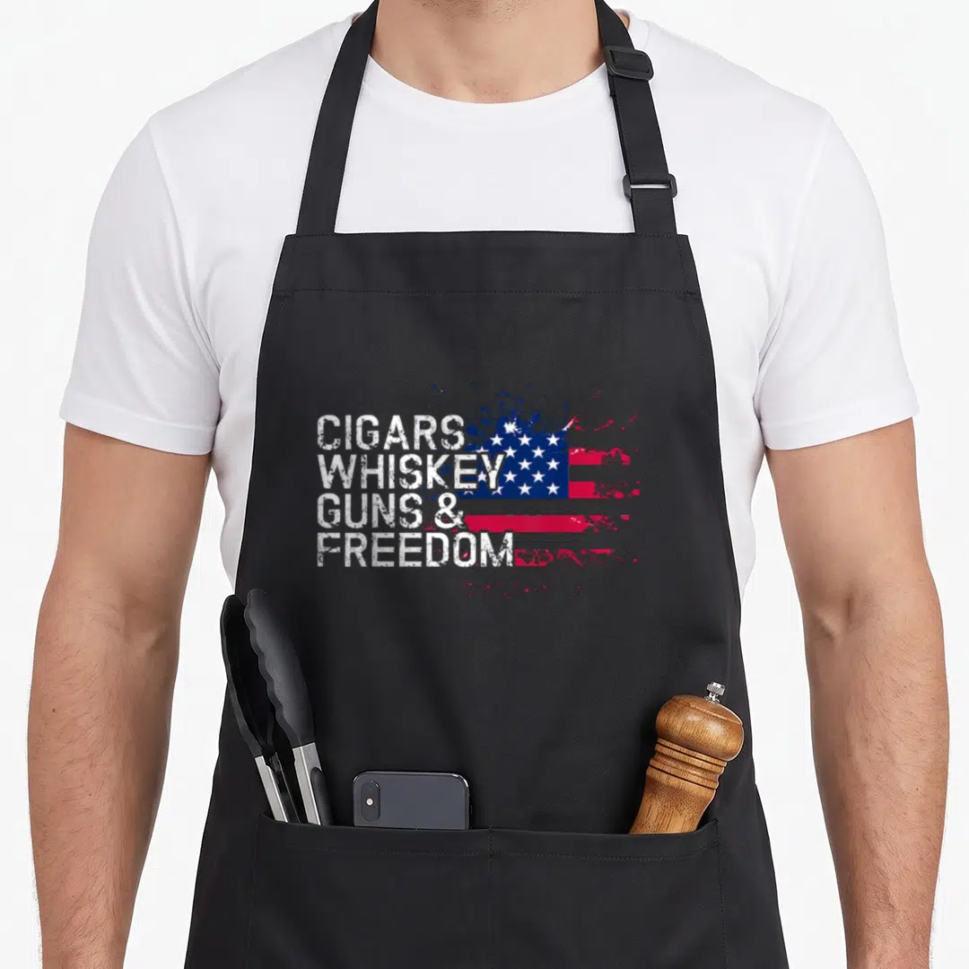 Freedom gun apron - Stacked white text Cigars Whiskey Guns Freedom over distressed splatter USA flag — shown on a black cotton canvas apron against a white background