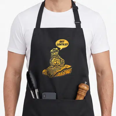 Thorn red tailed apron - Yellow cartoon toad on rock saying got lentils speech bubble — shown on a black cotton canvas apron against a white background