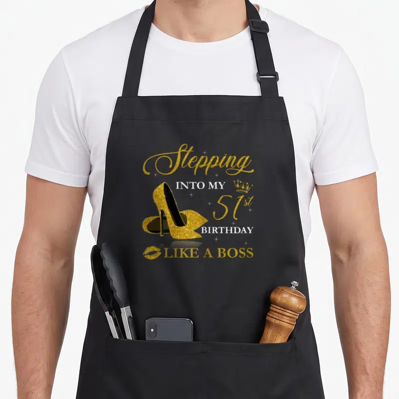 51st bbq boss apron - Gold glitter high heel shoe with Stepping Into My 51st Birthday Like A Boss text and crown icon — shown on a black cotton canvas apron against a white background