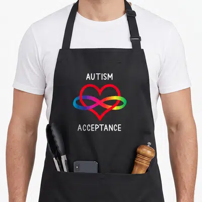 Autism pro apron - White autism acceptance text framing rainbow infinity symbol over red heart outline — shown on a black cotton canvas apron against a white background
