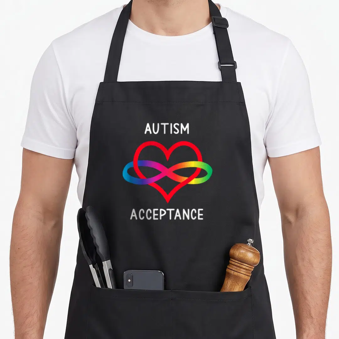 Autism pro apron - White autism acceptance text framing rainbow infinity symbol over red heart outline — shown on a black cotton canvas apron against a white background