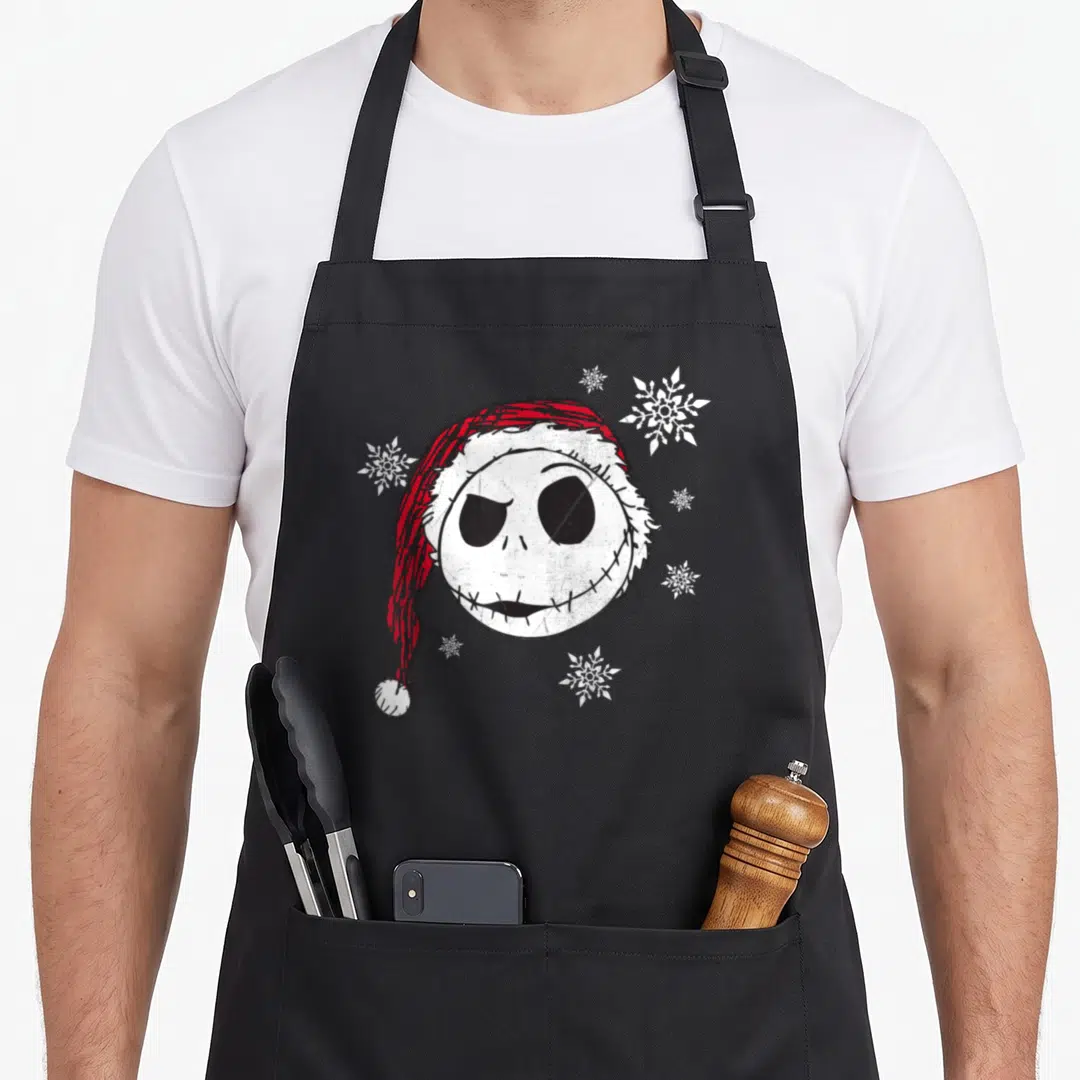 Funny snowflake holiday apron - Distressed Jack Skellington head in red Santa hat with scattered white snowflakes — shown on a black cotton canvas apron against a white background