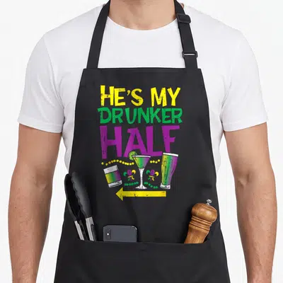 Hes mardi gras aprons - HE'S MY DRUNKER HALF text above three cocktails, beads, and fleur-de-lis symbols — shown on a black cotton canvas apron against a white background