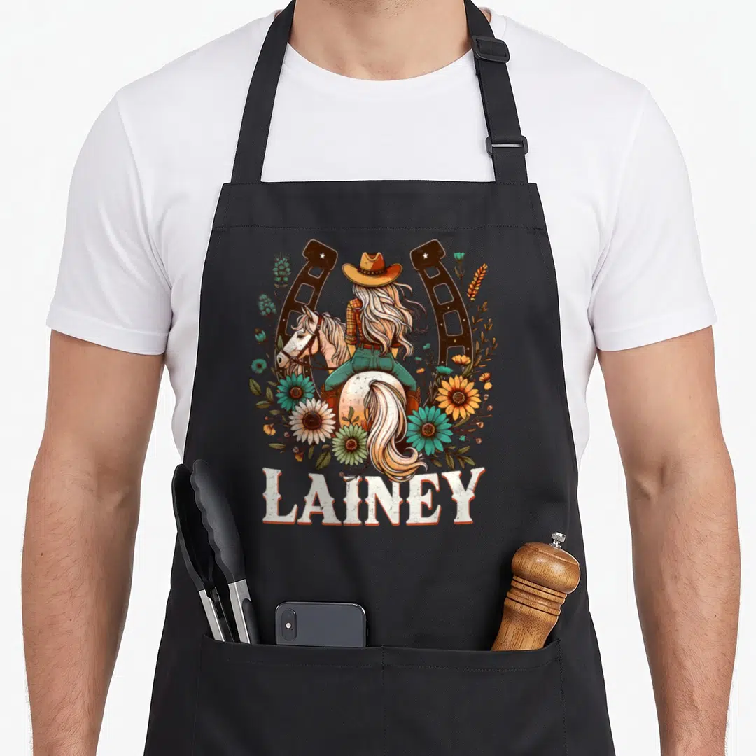 Lainey cowgirl aprons - Cowgirl on horse in horseshoe with flowers and name Lainey below — shown on a black cotton canvas apron against a white background