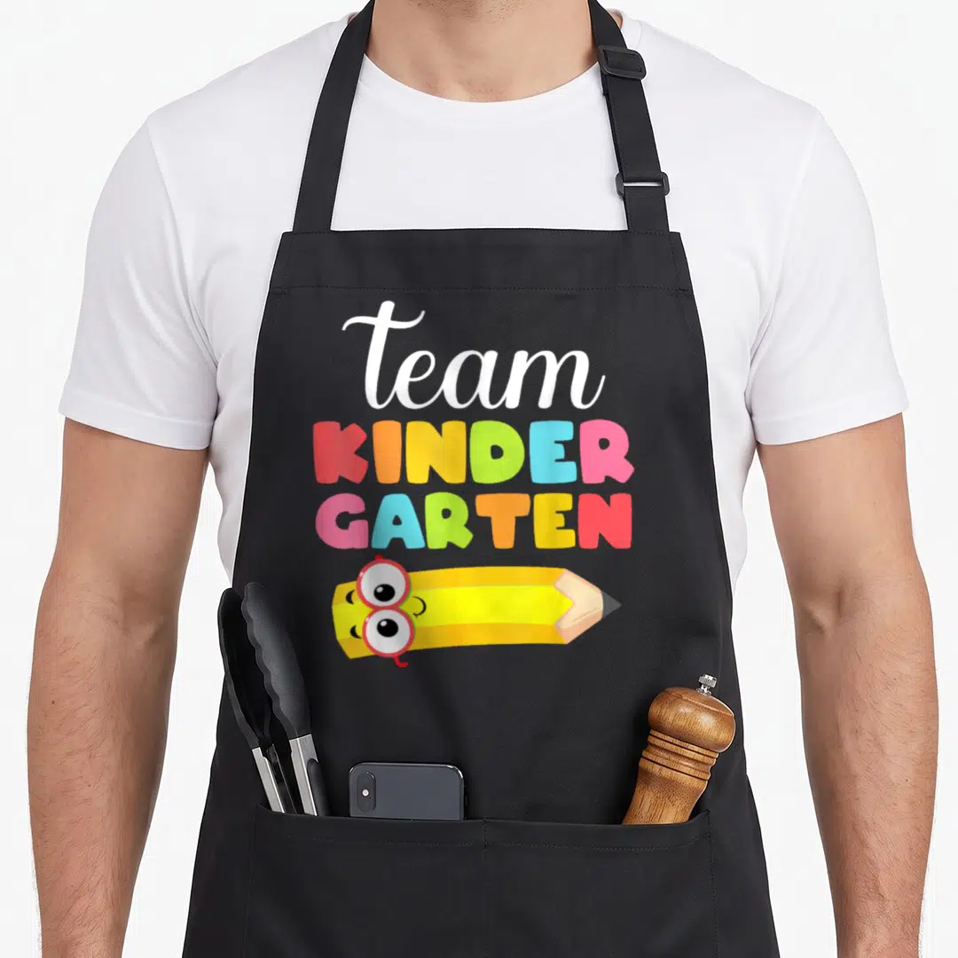 Funny student kindergarten teacher apron - Team Kinder Garten text above a yellow pencil wearing red glasses — shown on a black cotton canvas apron against a white background