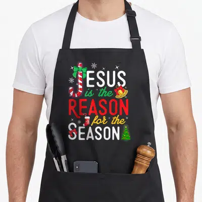 Reason womens christmas aprons - Stacked text JESUS is the REASON for the SEASON with candy cane J, bell, stocking — shown on a black cotton canvas apron against a white background