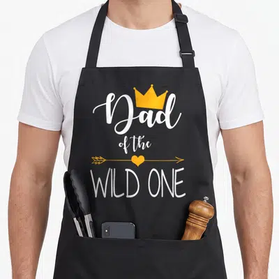 Cute wild birthday apron - Dad of the Wild One text with yellow crown and heart arrow — shown on a black cotton canvas apron against a white background