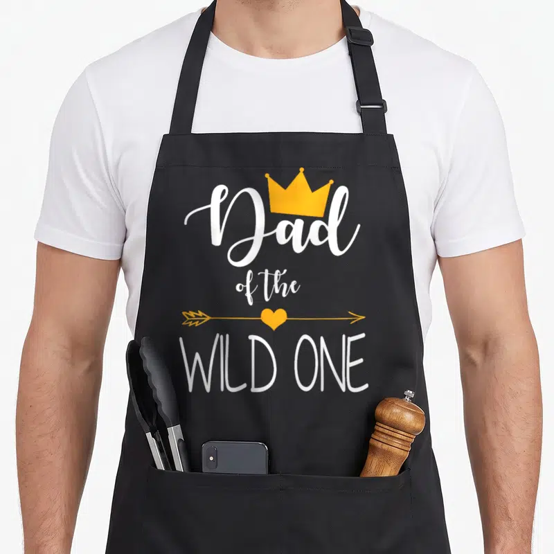 Cute wild birthday apron - Dad of the Wild One text with yellow crown and heart arrow — shown on a black cotton canvas apron against a white background