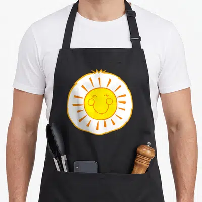 Funshine halloween apron - Cartoon sun with yellow face, white circle, and orange rays on canvas apron — shown on a black cotton canvas apron against a white background