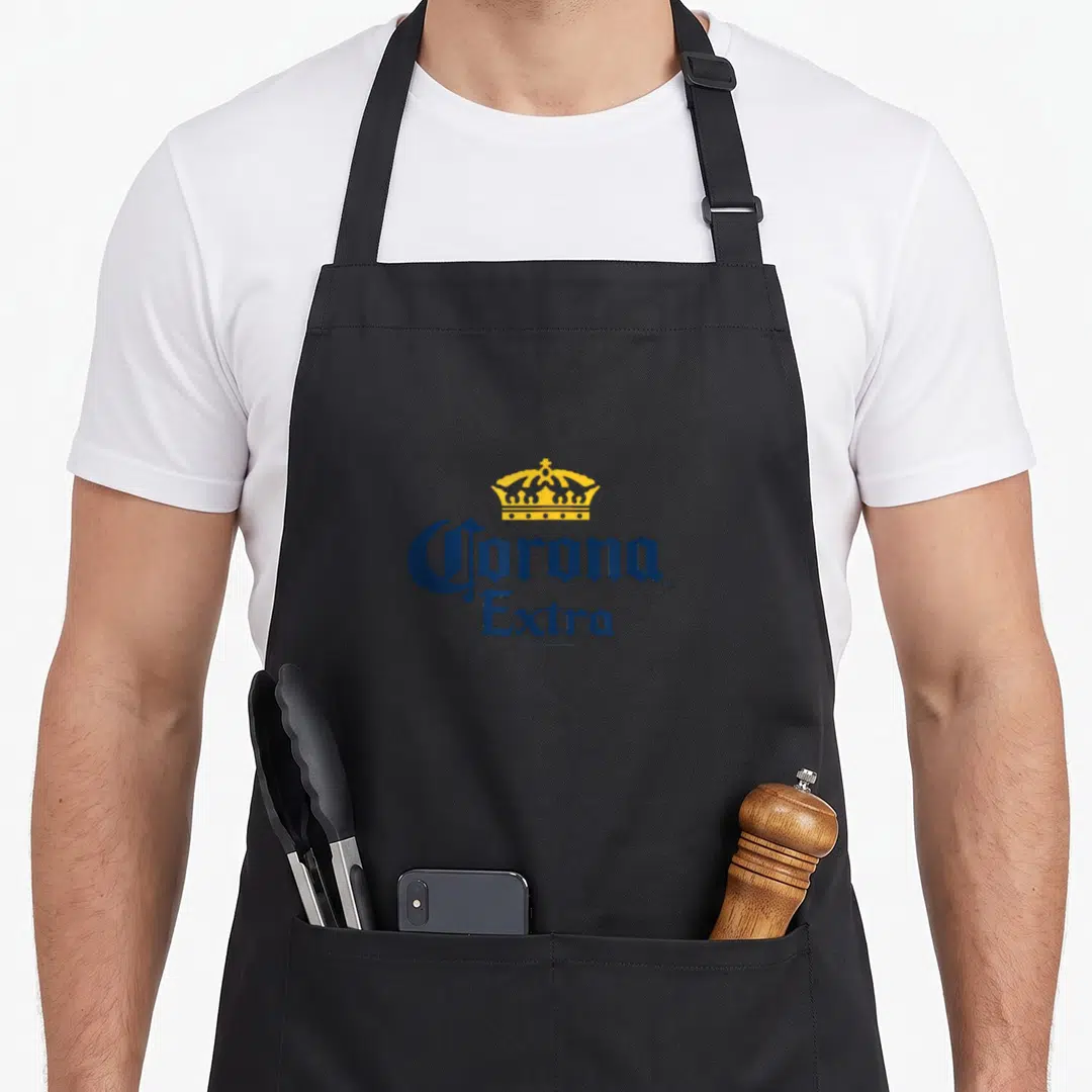 Corona an extra apron - Yellow crown centered above blue stacked Corona Extra typography — shown on a black cotton canvas apron against a white background