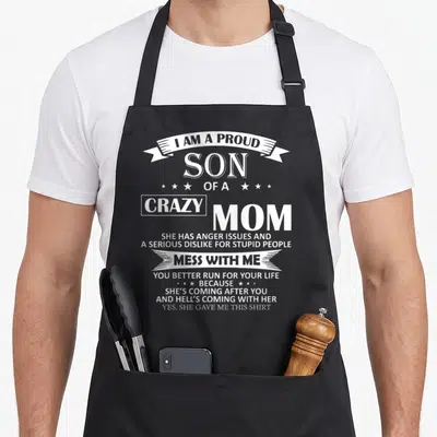 Proud mom and son aprons - White text reading I am a proud son of a crazy mom on black fabric — shown on a black cotton canvas apron against a white background