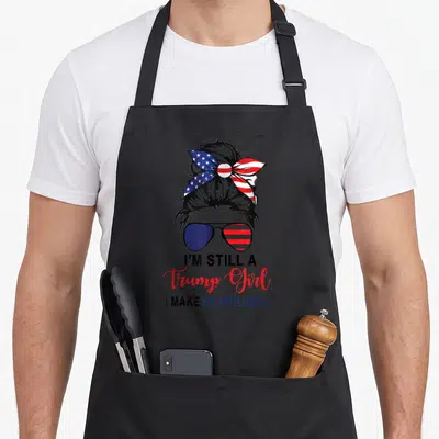 Trump patriotic apron - Woman's head silhouette with patriotic bow and sunglasses above I'm Still A Trump Girl text — shown on a black cotton canvas apron against a white background
