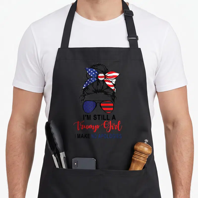 Trump patriotic apron - Woman's head silhouette with patriotic bow and sunglasses above I'm Still A Trump Girl text — shown on a black cotton canvas apron against a white background