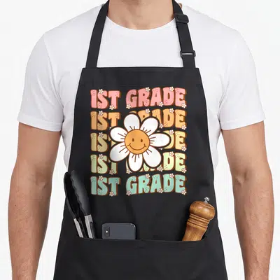 1st groovy grade apron - Five stacked 1ST GRADE lines with central smiling daisy and scattered small white flowers — shown on a black cotton canvas apron against a white background