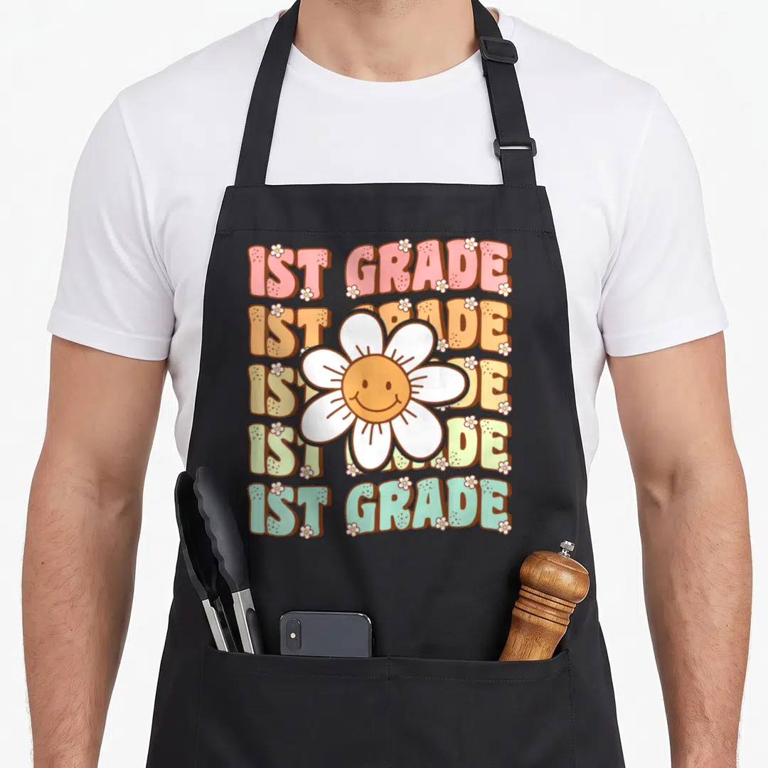 1st groovy grade apron - Five stacked 1ST GRADE lines with central smiling daisy and scattered small white flowers — shown on a black cotton canvas apron against a white background