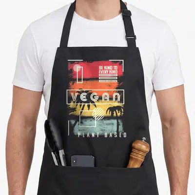 Kind plant based apron - Palm trees behind red orange yellow teal white stripes with BE KIND TO EVERY KIND VEGAN PLANT — shown on a black cotton canvas apron against a white background