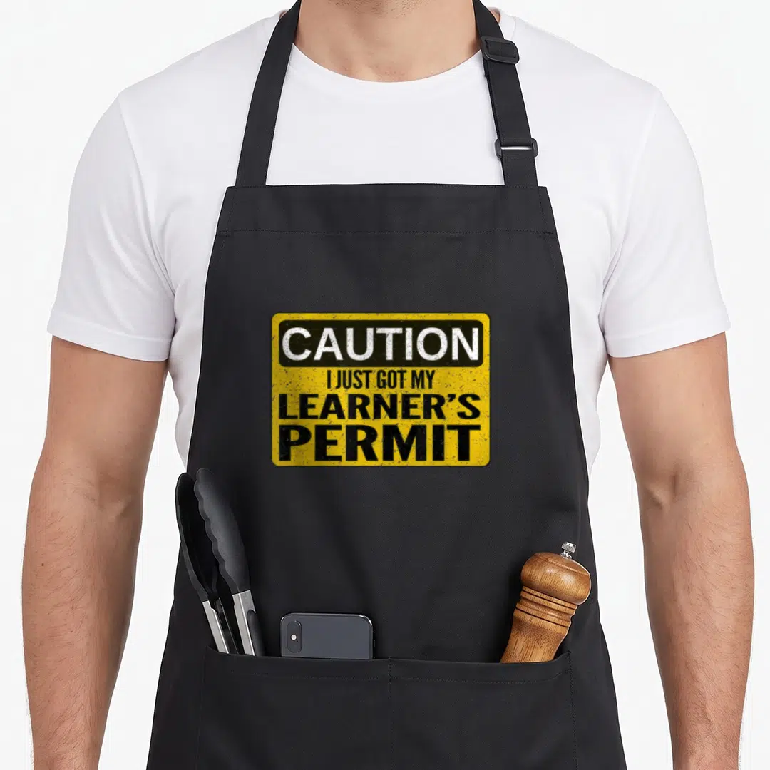 Learner permit apron - Yellow sign graphic with white and black text reading CAUTION I JUST GOT MY LEARNER'S PERMIT — shown on a black cotton canvas apron against a white background