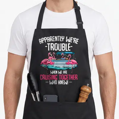 Apparently cruising lover apron - Four pink flamingos on a blue pontoon boat with teal water and white text — shown on a black cotton canvas apron against a white background