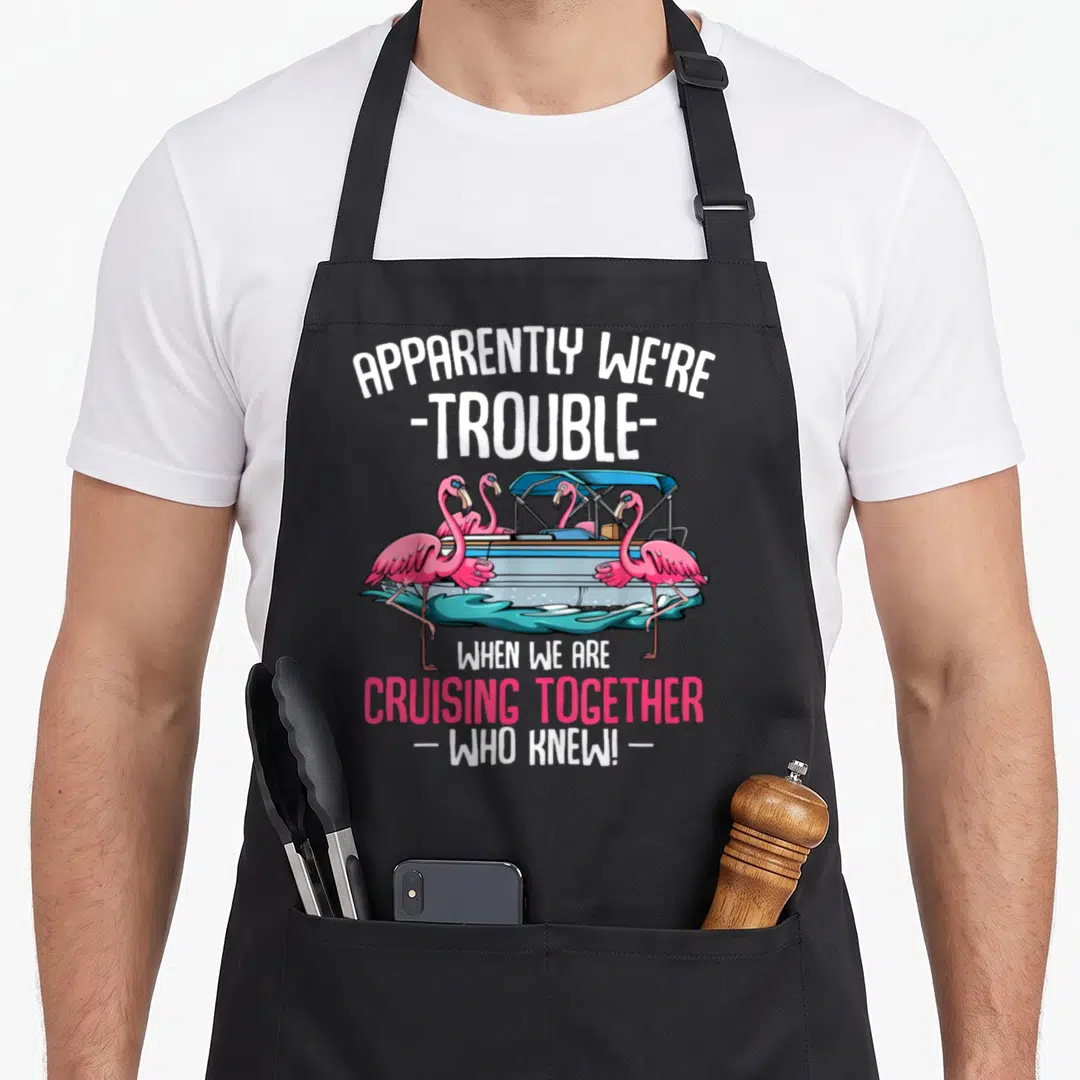 Apparently cruising lover apron - Four pink flamingos on a blue pontoon boat with teal water and white text — shown on a black cotton canvas apron against a white background