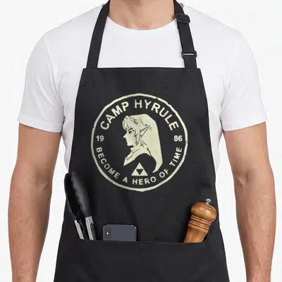 Link craft apron - Cream circular badge with Zelda profile, Triforce, and Camp Hyrule 1986 text — shown on a black cotton canvas apron against a white background