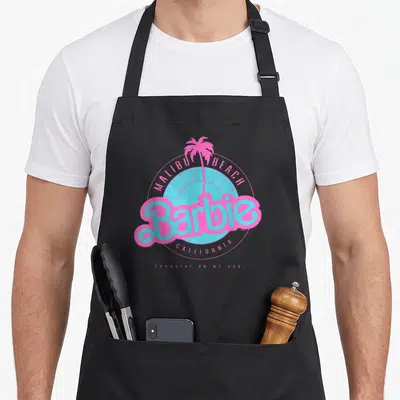 Beach california malibu apron - Pink Barbie logo over blue sun and palm tree, Malibu Beach California text, Sunshine — shown on a black cotton canvas apron against a white background