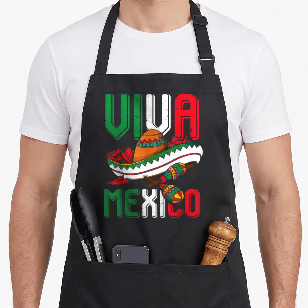 Mayo mexico apron - VIVA MEXICO text surrounding an orange sombrero, two maracas, and red chili peppers in green — shown on a black cotton canvas apron against a white background