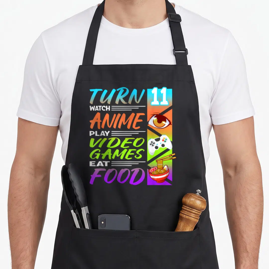 11th anime mom apron - Stacked text TURN 11 WATCH ANIME PLAY VIDEO GAMES EAT FOOD beside eye controller and ramen strip — shown on a black cotton canvas apron against a white background
