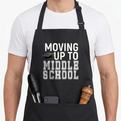 Moving diploma apron - Stacked white text reading Moving Up to Middle School with black graduation cap overlapping — shown on a black cotton canvas apron against a white background