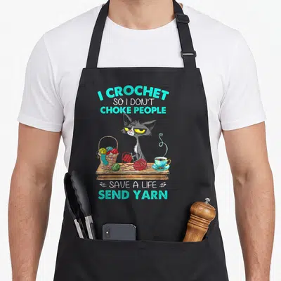 Send crochet apron - Grumpy grey cat at table with yarn basket and teal text reading send yarn — shown on a black cotton canvas apron against a white background